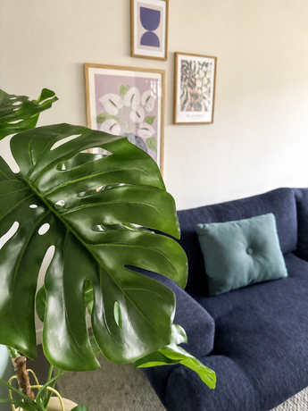 Im Vordergrund ist prominent eine Kunstpflanze Monstera 120 cm platziert. Dahinter steht ein dunkelblaues Sofa mit hellblauem Kissen vor einer beigen Wand mit drei gerahmten Kunstwerken.