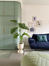 Ein modernes Wohnzimmer mit einem grünen Einbauschrank, einer künstlichen Pflanze Monstera 120 cm, einem blauen Sofa mit einem blaugrünen Kissen, abstrakter Kunst an der Wand und einem grünen Sitzpuff auf einem hellen Teppich.