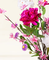Der künstliche Blumenstrauß Pink Reverie aus Seide besteht aus einer großen magentafarbenen Pfingstrose, kleinen rosa Blüten, violetten, gänseblümchenähnlichen Blüten und grünen Blättern in einer Vase – ein romantischer Strauß vor einem hellen Hintergrund.