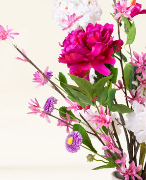 Der künstliche Blumenstrauß Pink Reverie aus Seide besteht aus einer großen magentafarbenen Pfingstrose, kleinen rosa Blüten, violetten, gänseblümchenähnlichen Blüten und grünen Blättern in einer Vase – ein romantischer Strauß vor einem hellen Hintergrund.