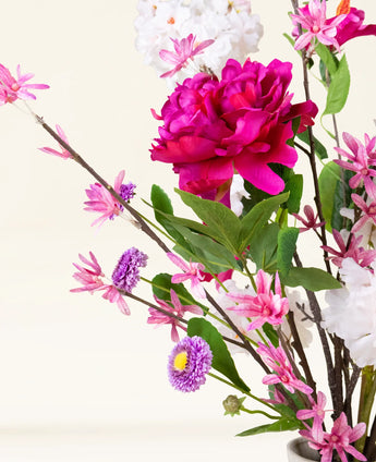 Der künstliche Blumenstrauß Pink Reverie aus Seide besteht aus einer großen magentafarbenen Pfingstrose, kleinen rosa Blüten, violetten, gänseblümchenähnlichen Blüten und grünen Blättern in einer Vase – ein romantischer Strauß vor einem hellen Hintergrund.