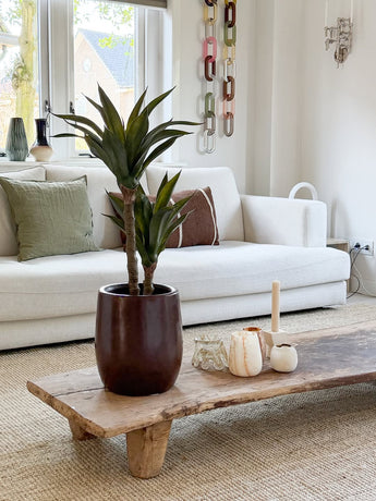 Ein modernes Wohnzimmer mit einem weißen Sofa, grünen und braunen Kissen, einem mit Kerzen und der künstlichen Agavenpflanze 76 cm dekorierten Couchtisch aus Holz sowie einer Wanddekoration aus Kettengeflecht. Durch die Fenster hinter dem Sofa fällt natürliches Licht ein.