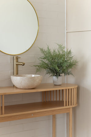Ein minimalistisches Badezimmermöbel verfügt über einen runden Spiegel, einen goldenen Wasserhahn, ein Steinwaschbecken und die künstliche Spargelpflanze (43 cm) in einem dekorativen Topf auf einer hellen Holzarbeitsplatte vor weiß gefliesten Wänden.