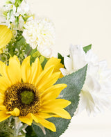Eine Nahaufnahme des künstlichen Seidenblumenstraußes Sunny Whisper mit einer leuchtend gelben Sonnenblume, weißen Dahlien und grünen Blättern vor einem hellen, neutralen Hintergrund.