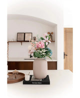 Die Vase Artic Beige Ø21 x H29 cm, gefüllt mit rosa und blauen Kunstblumen, steht auf einem schwarzen Buch auf einer weißen Arbeitsplatte. Sanftes Licht betont die minimalistische Szene mit dekorativen Regalen und einer Holztür im Hintergrund.