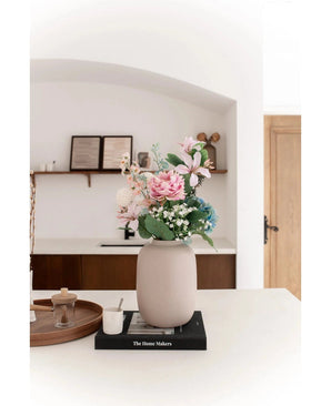 Die Vase Artic Beige Ø21 x H29 cm, gefüllt mit rosa und blauen Kunstblumen, steht auf einem schwarzen Buch auf einer weißen Arbeitsplatte. Sanftes Licht betont die minimalistische Szene mit dekorativen Regalen und einer Holztür im Hintergrund.