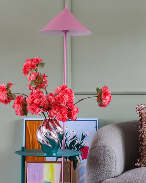 Eine rosa Lampe, eine gestreifte Vase mit dem Silk Artificial Flower Bouquet Cherry Blossom Coral, farbenfrohe Kunstwerke und ein grauer Stuhl stehen vor einer hellgrünen Wand und schaffen eine gemütliche, lebendige Ecke.