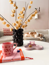 Ein gemütliches Wohnzimmer mit einem beigen Sofa, einer schlafenden Katze, einer skulpturalen schwarzen Vase mit der Kunstblume Physalis Lampion 90 cm braun, einer rosa Tasse „LOVE“, einem Buch, einer Kerze und einem Keks auf dem Couchtisch.