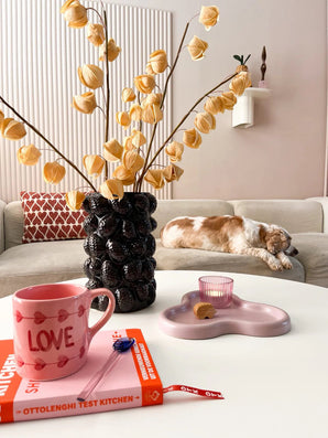 Ein gemütliches Wohnzimmer mit einem beigen Sofa, einer schlafenden Katze, einer skulpturalen schwarzen Vase mit der Kunstblume Physalis Lampion 90 cm braun, einer rosa Tasse „LOVE“, einem Buch, einer Kerze und einem Keks auf dem Couchtisch.